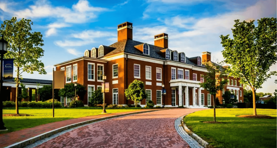Elegant brick building with white columns, surrounded by lush greenery and a winding brick pathway, under a blue sky with scattered clouds.