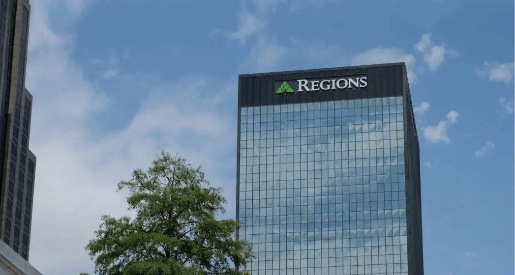 Regions Bank building with reflective glass windows, topped with a green logo, adjacent to green trees and a blue sky.