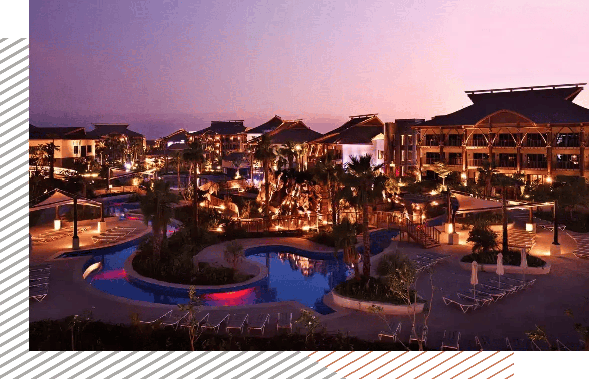 Luxury resort at sunset with illuminated pools, palm trees, and lounge areas.