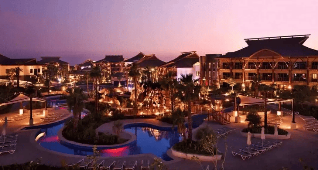 A luxurious resort at twilight, featuring illuminated pools, palm trees, and elegant architecture against a colorful sunset sky.