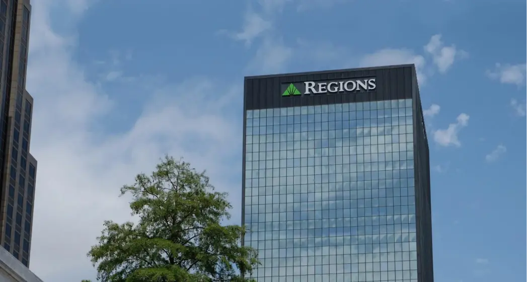 Regions Bank building with reflective glass windows, topped with a green logo, adjacent to green trees and a blue sky.