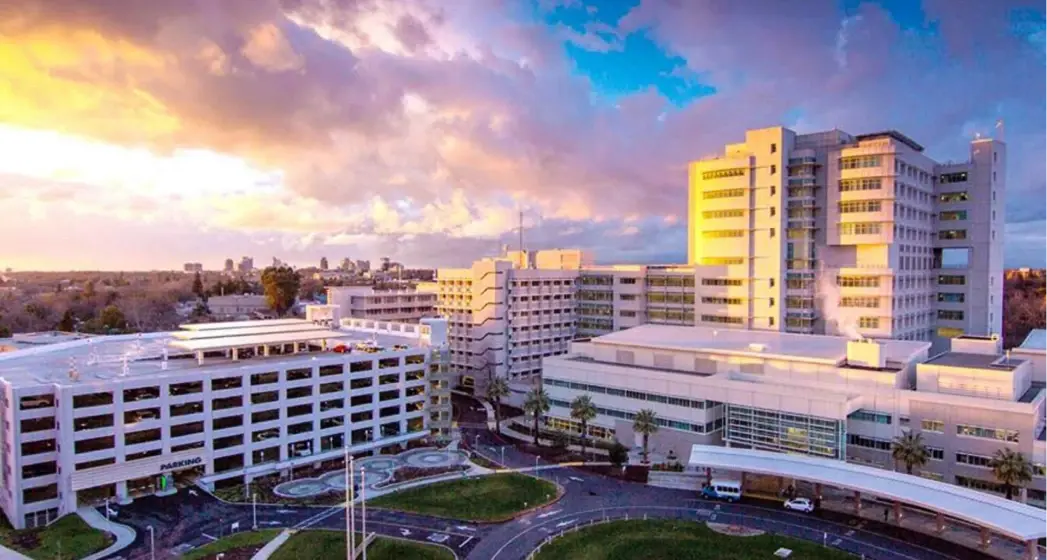 Aerial view of a modern multistory hospital complex under a colorful sunset, with a parking structure and landscaped areas.