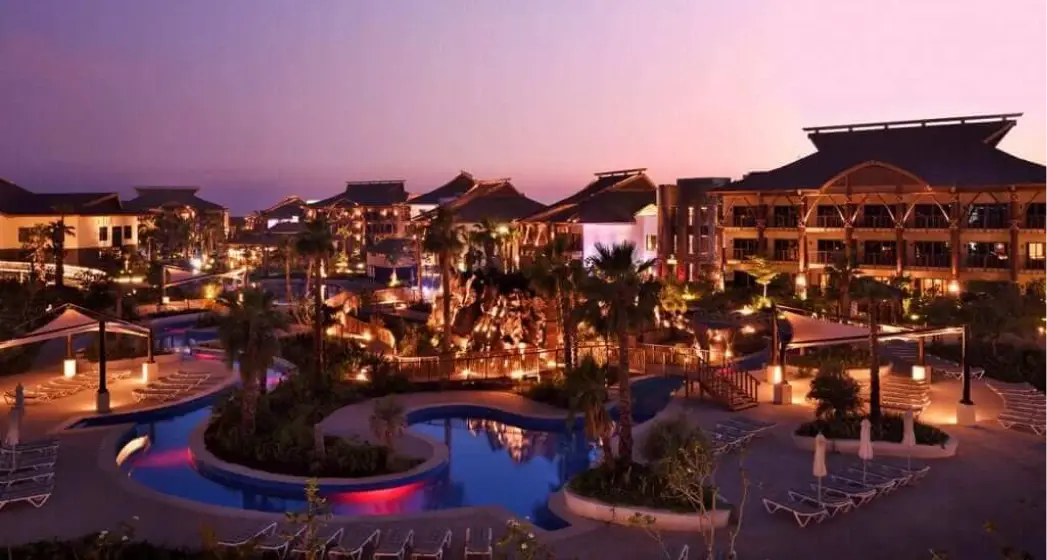 A luxurious resort at twilight, featuring illuminated pools, palm trees, and elegant architecture against a colorful sunset sky.